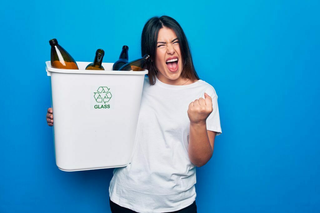 Young beautiful woman recycling glass bottles on wastebasket to care environment screaming proud, celebrating victory and success very excited with raised arm