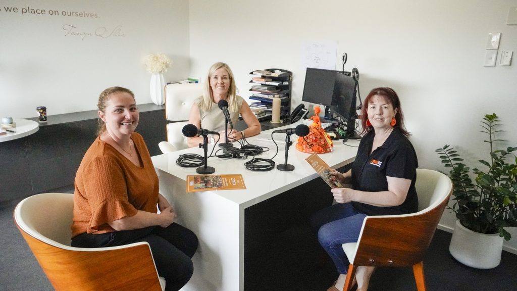 Staci Rae, Tanya O'Shea, and Jo Leveritt  recording a podcast session in a modern office setup with microphones on the table, surrounded by plants and motivational quotes on the wall.